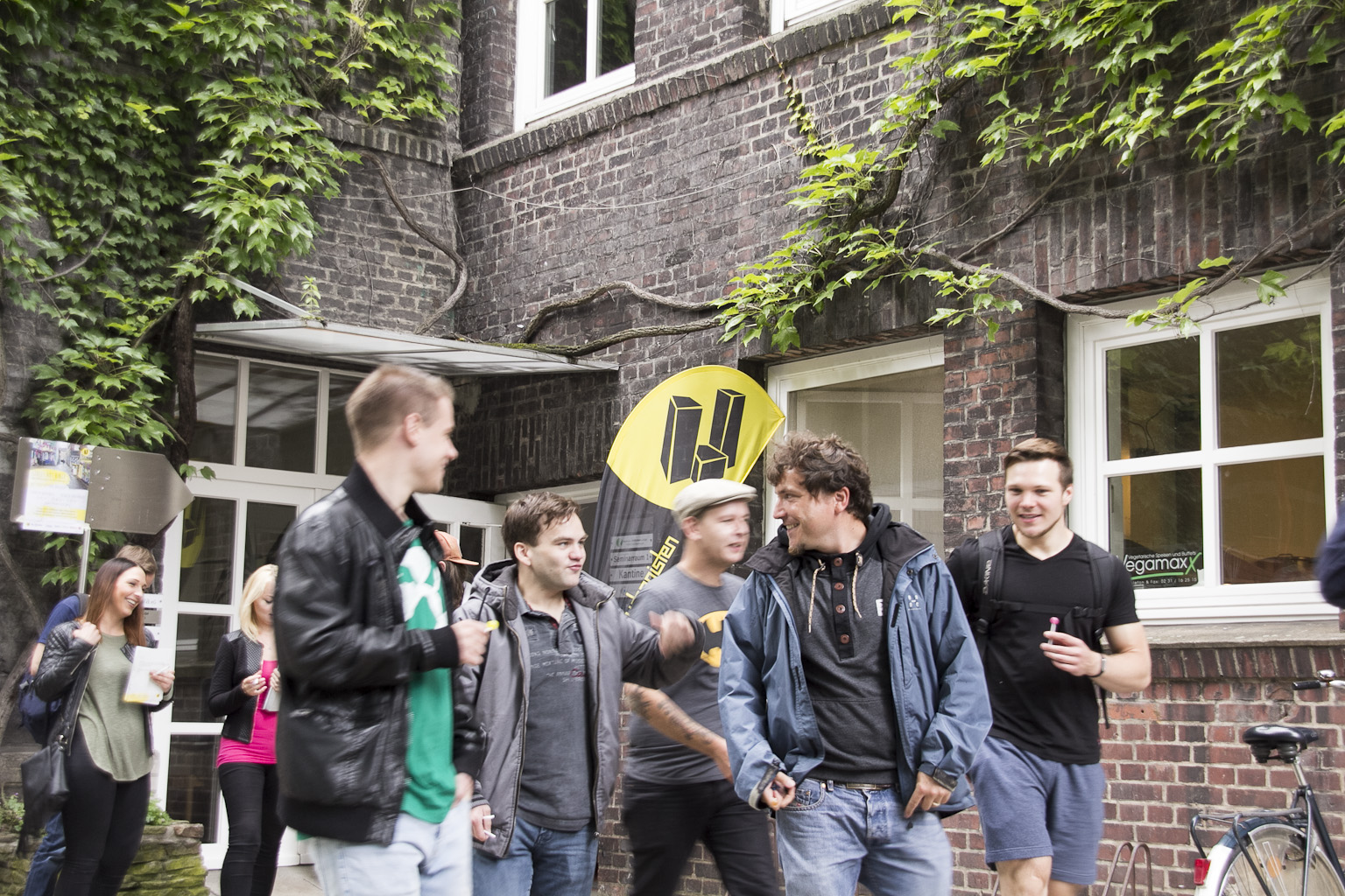 Gruppe junger Menschen steht vor einem Backsteingebäude mit Efeu an der Fassade und einem gelben Werbefahnen