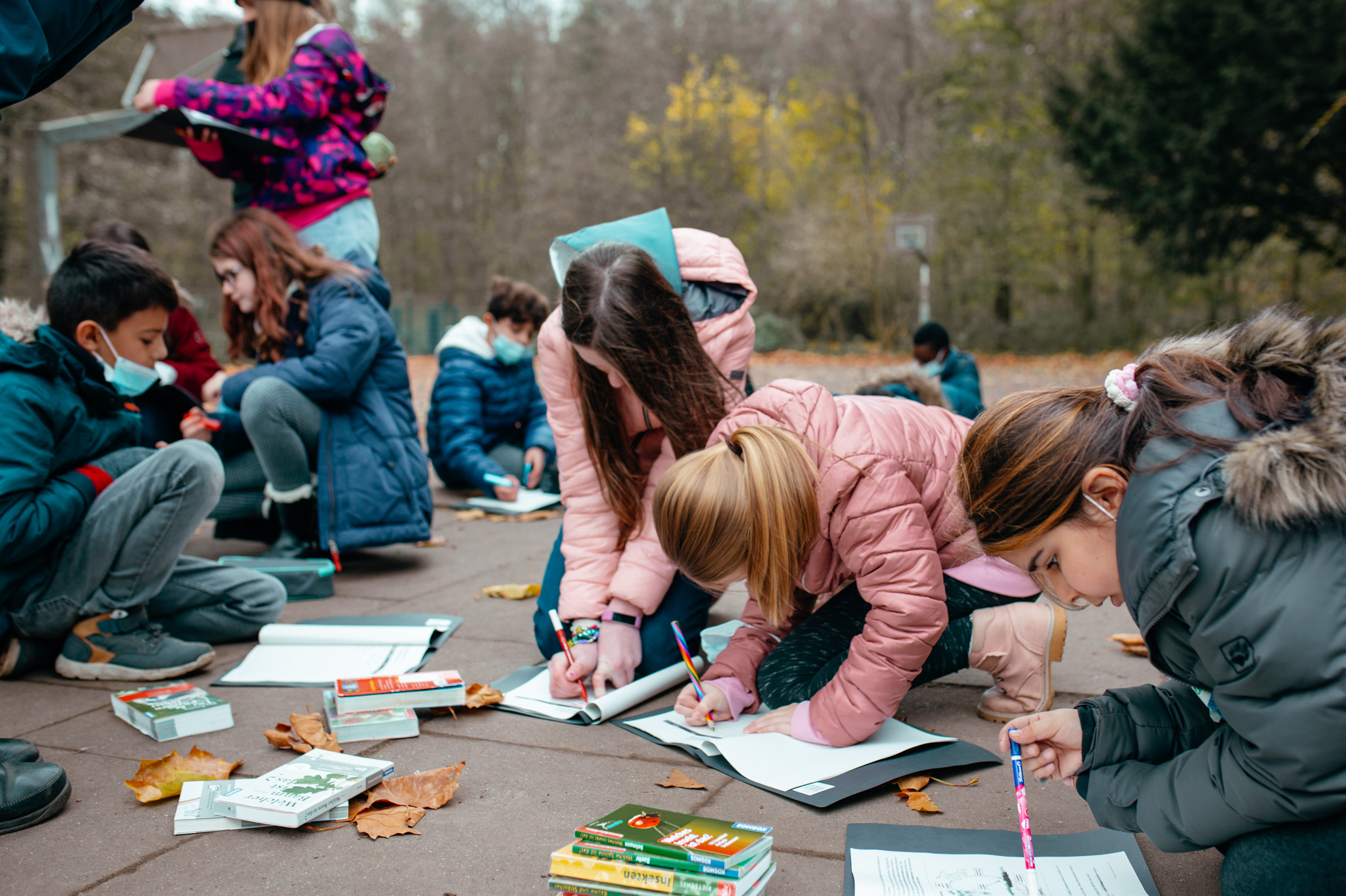 Kinder sitzen und knien auf dem Boden im Freien und schreiben oder zeichnen in Hefte, umgeben von Büchern und Herbstlaub